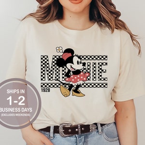 May include: A white t-shirt with a black and white checkered design featuring a cartoon image of Minnie Mouse in a red dress and yellow shoes. The text "Minnie" is written in black letters on the checkered background. The year "1928" is printed below the checkered design.
