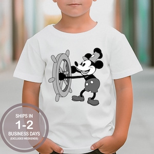 May include: A white t-shirt with a black and white image of Mickey Mouse as a sailor holding a ship's wheel. The text "SHIPS IN 1-2 BUSINESS DAYS (EXCLUDES WEEKENDS)" is printed on the bottom of the shirt.