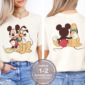 May include: A cream-colored t-shirt with a cartoon image of Mickey Mouse and Pluto. Mickey Mouse is wearing red shorts and Pluto is wearing a green collar. The image is on the front of the shirt. The back of the shirt has the same image but Mickey Mouse and Pluto are facing the opposite direction.