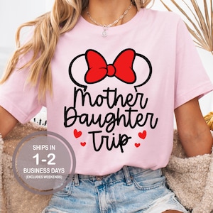 Puede incluir: Camiseta rosa con un lazo rojo y el texto "Mother Daughter Trip" en negro con corazones rojos.