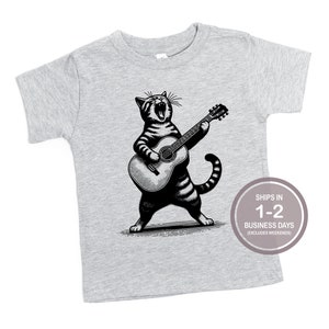 May include: A gray short-sleeved t-shirt with a black and white illustration of a cat playing a guitar. The cat is singing and has a striped tail.
