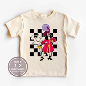 May include: A cream-colored short-sleeve t-shirt featuring a graphic of Captain Hook in a red coat and purple hat, holding a sword. Behind the character is a black and white checkered square pattern with the text Captain Hook in script font.