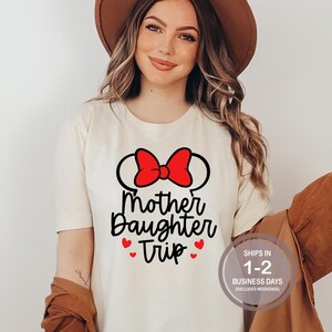 Puede incluir: Camiseta blanca con un lazo rojo y texto negro que dice "Mother Daughter Trip" con tres corazones rojos.