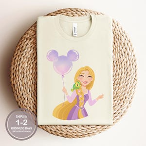 May include: A cream-colored t-shirt featuring a graphic of a long-haired blonde character in a purple dress holding a purple Mickey Mouse-shaped balloon with a small green chameleon perched on their braided hair, with a tag reading CANVAS.