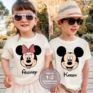 May include: Two children wearing cream-colored t-shirts with Disney character graphics. One shirt features Minnie Mouse with a pink bow and the name "Audrey". The other shirt has Mickey Mouse and the name "Kasen". Both shirts have black graphics.