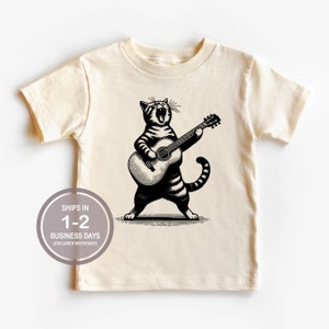 May include: A cream colored t-shirt with a black and white illustration of a cat playing a guitar. The cat is singing and has a striped pattern.