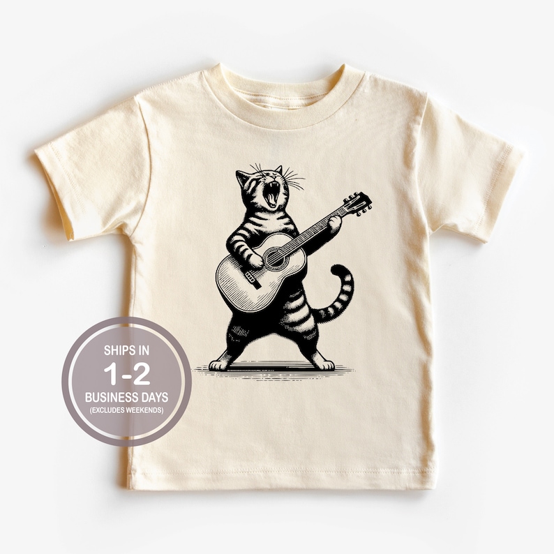 May include: A cream colored t-shirt with a black and white illustration of a cat playing a guitar. The cat is singing and has a striped pattern.