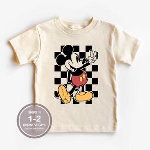 Puede incluir: Camiseta color crema con un gr&aacute;fico de Mickey Mouse haciendo el signo de la paz frente a un fondo a cuadros blanco y negro. Mickey lleva pantalones cortos rojos y zapatos amarillos. La camiseta tiene el texto "SHIPS IN 1-2 BUSINESS DAYS (EXCLUDES WEEKENDS)" en un c&iacute;rculo.