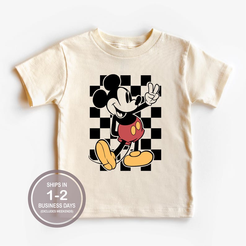 Puede incluir: Camiseta color crema con un gr&aacute;fico de Mickey Mouse haciendo el signo de la paz frente a un fondo a cuadros blanco y negro. Mickey lleva pantalones cortos rojos y zapatos amarillos. La camiseta tiene el texto "SHIPS IN 1-2 BUSINESS DAYS (EXCLUDES WEEKENDS)" en un c&iacute;rculo.