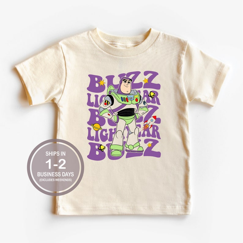 K&ouml;nnte beinhalten: Ein hellbeiges T-Shirt mit einer lila und wei&szlig;en Grafik von Buzz Lightyear aus der Toy Story-Reihe. Die Grafik zeigt Buzz Lightyear stehend mit verschr&auml;nkten Armen und dem Text "BUZZ LIGHTYEAR" in einem Retro-Stil wiederholt.