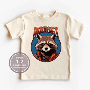 May include: A cream colored t-shirt with a cartoon image of Rocket Raccoon from Guardians of the Galaxy. The image is surrounded by a blue circle with the word "ROCKET" in orange letters.