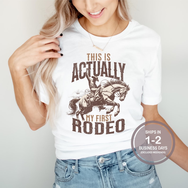 May include: White t-shirt with a brown graphic of a cowboy riding a horse. The text "This is Actually My First Rodeo" is printed on the shirt.