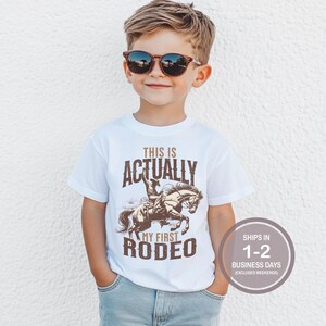 May include: White t-shirt with a brown graphic of a cowboy riding a horse. The text on the shirt reads "This is Actually My First Rodeo".