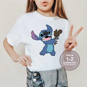 May include: White t-shirt with a blue Stitch character holding a Mickey Mouse ice cream. The text "SHIPS IN 1-2 BUSINESS DAYS (EXCLUDES WEEKENDS)" is on a white circle.