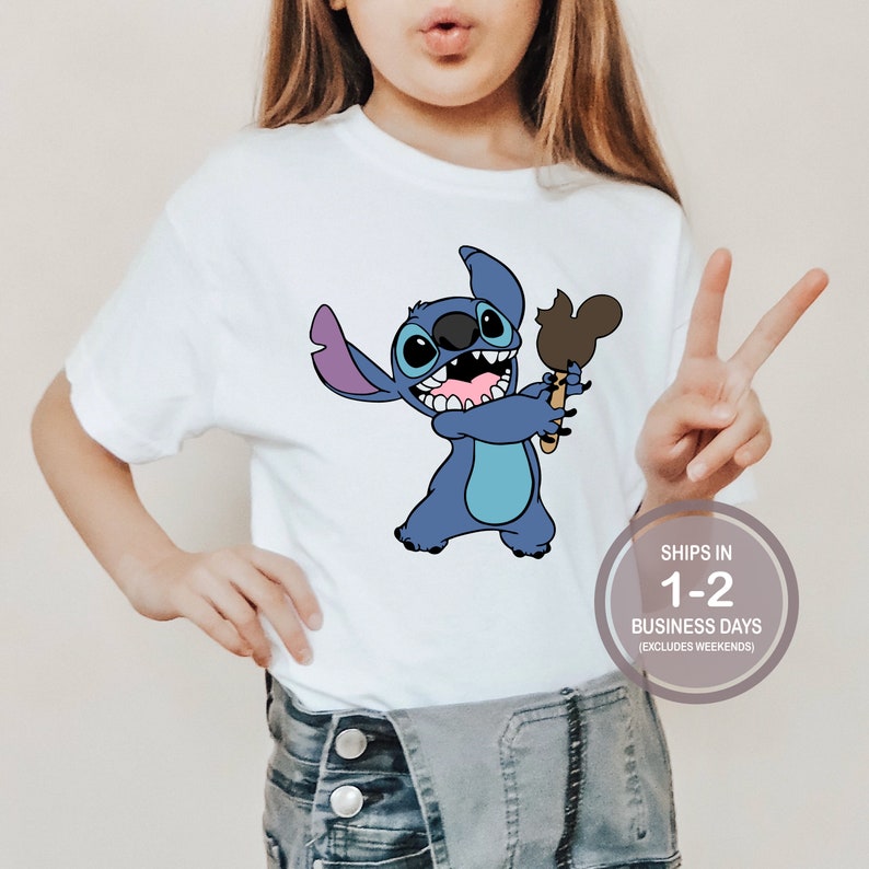 May include: White t-shirt with a blue Stitch character holding a Mickey Mouse ice cream. The text "SHIPS IN 1-2 BUSINESS DAYS (EXCLUDES WEEKENDS)" is on a white circle.