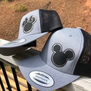 May include: Two gray and black baseball caps with a black Mickey Mouse silhouette on one and a black Minnie Mouse silhouette on the other. The caps have a black mesh back and a silver label that reads "The Authentic Snapback Yupoong Since 1974".