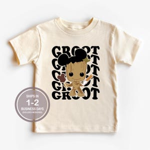 May include: A beige t-shirt with a black graphic of Groot from Guardians of the Galaxy wearing Mickey Mouse ears and holding a Mickey Mouse ice cream bar. The text "GROOT" is repeated in black letters on the shirt.