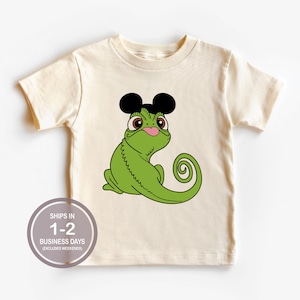 May include: A cream-colored short-sleeve t-shirt featuring a graphic illustration of a green chameleon with large brown eyes and a pink tongue sticking out. The chameleon is wearing black mouse-ear headwear on its head and has a curled tail.