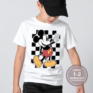 Puede incluir: Camiseta blanca con un personaje de dibujos animados de Mickey Mouse haciendo el signo de la paz. Mickey Mouse est&aacute; representado en sus colores cl&aacute;sicos: negro, rojo y amarillo. El fondo es un patr&oacute;n de cuadros blancos y negros.