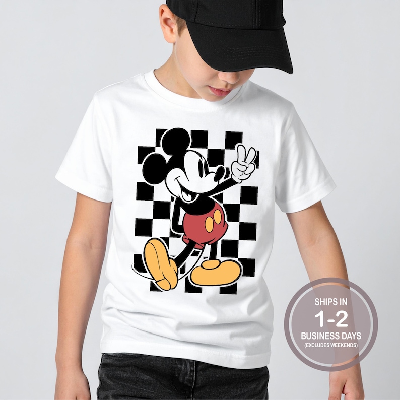 Puede incluir: Camiseta blanca con un personaje de dibujos animados de Mickey Mouse haciendo el signo de la paz. Mickey Mouse est&aacute; representado en sus colores cl&aacute;sicos: negro, rojo y amarillo. El fondo es un patr&oacute;n de cuadros blancos y negros.