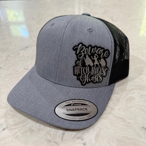 May include: Gray snapback hat with a black mesh back. The hat features the text "Beware of Hitch Hiking Ghosts" in a stylized font, along with a graphic of three ghost figures. The hat has a curved brim and a snap closure.