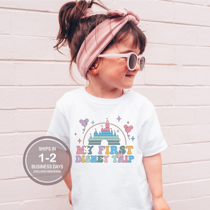 Może przedstawiać: Biała koszulka z kolorową grafiką zamku Kopciuszka i tekstem "My First Disney Trip".