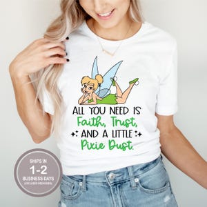 May include: White t-shirt featuring a cartoon fairy with green and blue accents. The text reads "ALL YOU NEED IS Faith, Trust, AND A LITTLE Pixie Dust." The shirt is paired with blue jeans.