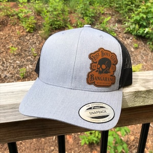 May include: A gray and black baseball cap with a brown leather patch featuring a skull and the text "Lost Boys Club 19 53 Bangarang! Neverland". The cap has a black mesh back and a YUPOONG SNAPBACK label.