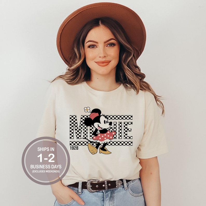 May include: A white t-shirt with a black and white checkered design featuring a cartoon image of Minnie Mouse in a red dress and yellow shoes. The text "Minnie" is written in black letters on the checkered background. The year "1928" is printed below the checkered design.