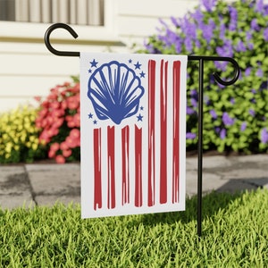 May include: A white garden flag with a red, white, and blue American flag design. A blue seashell is in the center of the flag with white stars around it.
