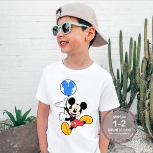 Puede incluir: Camiseta blanca con un gr&aacute;fico de Mickey Mouse sosteniendo un globo azul con orejas de Mickey Mouse. El texto "SHIPS IN 1-2 BUSINESS DAYS (EXCLUDES WEEKENDS)" est&aacute; en la esquina inferior derecha.