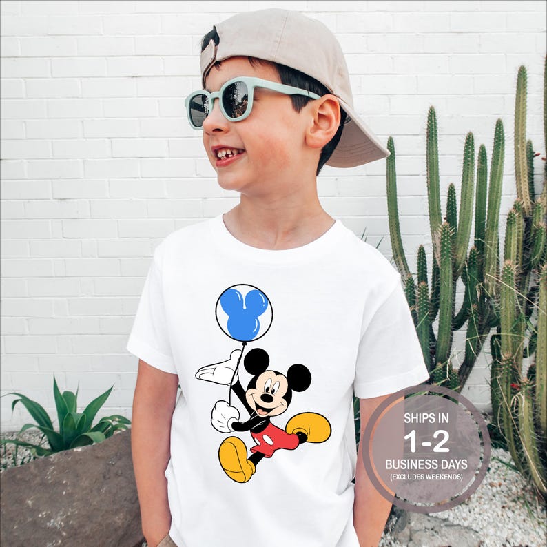 Puede incluir: Camiseta blanca con un gr&aacute;fico de Mickey Mouse sosteniendo un globo azul con orejas de Mickey Mouse. El texto "SHIPS IN 1-2 BUSINESS DAYS (EXCLUDES WEEKENDS)" est&aacute; en la esquina inferior derecha.
