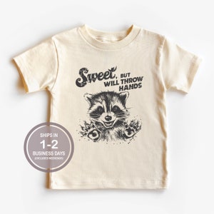 以下が含まれることがあります： クリーム色のTシャツに、黒と白のラッコのイラストと「Sweet, but will throw hands」というテキストがプリントされています。