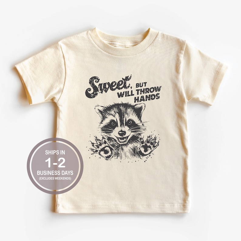 以下が含まれることがあります： クリーム色のTシャツに、黒と白のラッコのイラストと「Sweet, but will throw hands」というテキストがプリントされています。