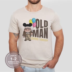 May include: A beige short-sleeve t-shirt featuring a cartoon illustration of an elderly character wearing black mouse ears and holding three colorful balloons. The text OLD MAN COMING THROUGH is printed in large brown and yellow block letters.