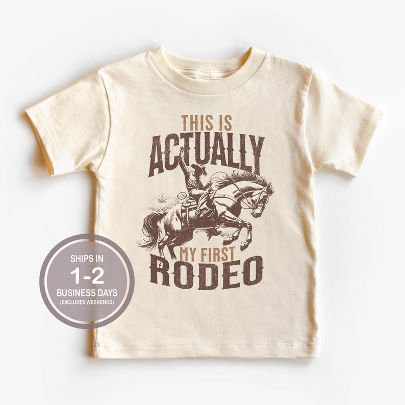 May include: A beige t-shirt with a brown graphic of a cowboy riding a horse. The text "This is Actually My First Rodeo" is printed on the shirt.