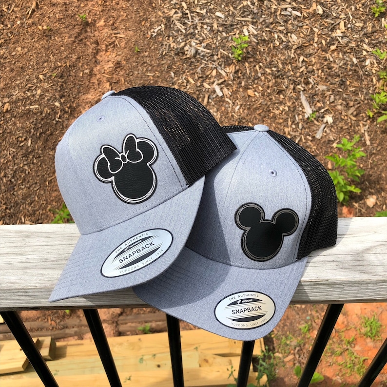 May include: Two gray and black baseball caps with a black Mickey Mouse and Minnie Mouse design on the front. The caps have a black mesh back and a white "The Authentic Snapback" label on the side.