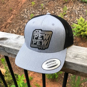 May include: A gray and black trucker hat with a silver patch that says "PEW PEW PEW" in a retro font. The hat has a black mesh back and a gray front. The hat is a Yupoong brand snapback.