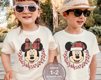 Könnte beinhalten: Zwei Kinder tragen weiße T-Shirts mit einem weihnachtlichen Mickey-Mouse-Motiv. Das Shirt des Mädchens zeigt Minnie Mouse mit einer rot-schwarzen Karobinde, während das Shirt des Jungen Mickey Mouse mit einem Weihnachtsmann-Hut zeigt. Beide Shirts zeigen einen rot-weißen karierten Kranz mit bunten Lichtern.