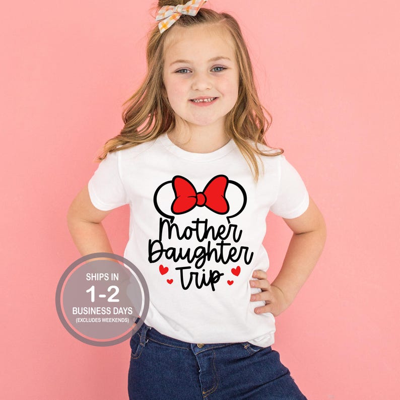 Puede incluir: Camiseta blanca con un lazo rojo y un gr&aacute;fico de orejas de rat&oacute;n. El texto "Mother Daughter Trip" est&aacute; impreso en negro debajo del gr&aacute;fico. Hay cuatro corazones rojos alrededor del texto.
