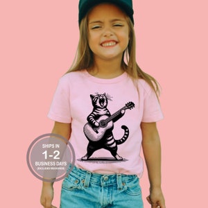 May include: A pink t-shirt with a black and white illustration of a cat playing a guitar. The cat is singing and has a striped pattern. The text "Ships in 1-2 Business Days (Excludes Weekends)" is on a white circle in the lower left corner of the image.