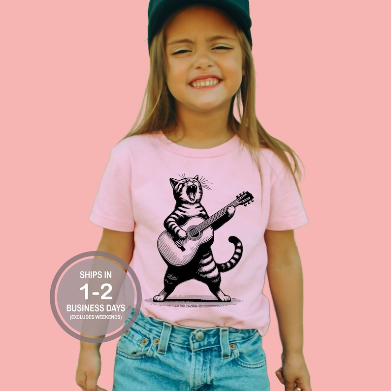 May include: A pink t-shirt with a black and white illustration of a cat playing a guitar. The cat is singing and has a striped pattern. The text "Ships in 1-2 Business Days (Excludes Weekends)" is on a white circle in the lower left corner of the image.