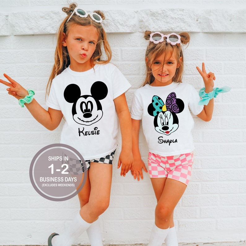 Pode incluir: Duas camisetas brancas com um Mickey Mouse preto e branco e um Minnie Mouse colorido. As camisetas t&ecirc;m os nomes "KelsIe" e "Shayla" impressos nelas.