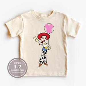 May include: A cream-coloured t-shirt with a cartoon illustration of a character with a red hat and a pink balloon. The character is wearing a white shirt, blue belt, and black and white trousers. The shirt has the text "SHIPS IN 1-2 BUSINESS DAYS (EXCLUDES WEEKENDS)" in a circle.