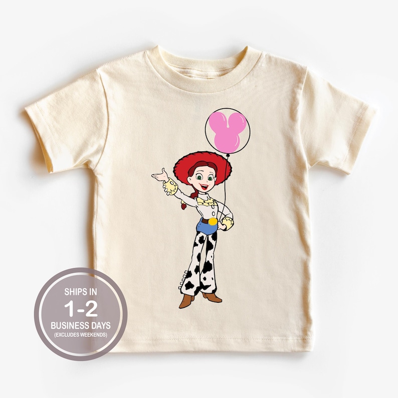May include: A cream-coloured t-shirt with a cartoon illustration of a character with a red hat and a pink balloon. The character is wearing a white shirt, blue belt, and black and white trousers. The shirt has the text "SHIPS IN 1-2 BUSINESS DAYS (EXCLUDES WEEKENDS)" in a circle.