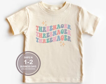 May include: A light beige short-sleeved t-shirt with the text "THREEENAGER" repeated three times in a rainbow color scheme with a retro font. The text is stacked vertically and has a sparkle effect.
