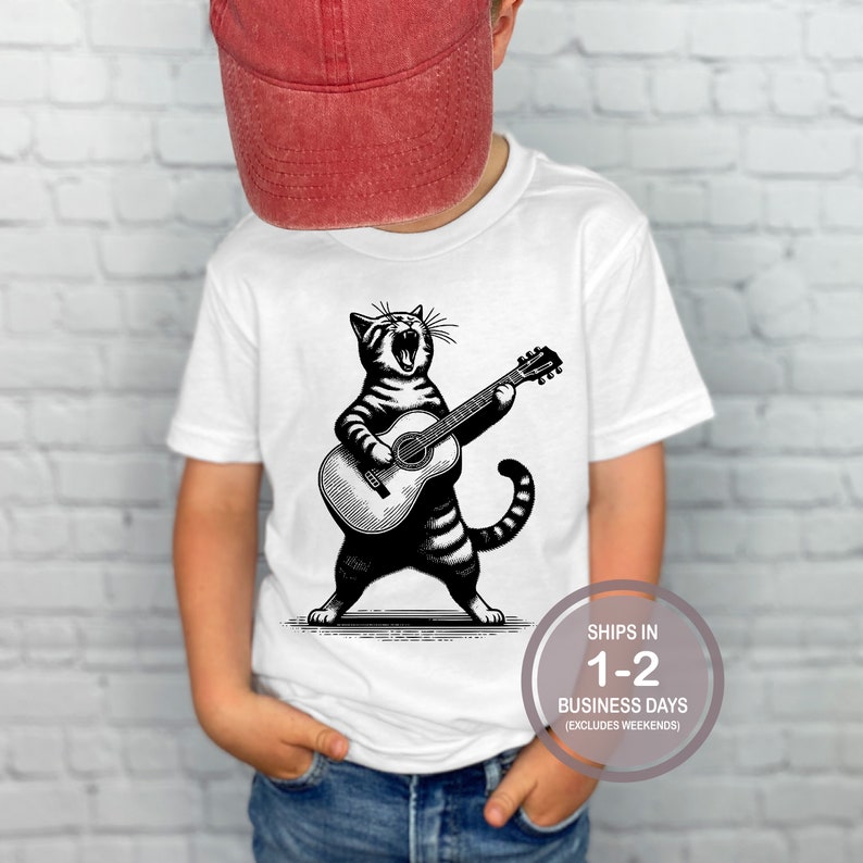 May include: White t-shirt with a black and white illustration of a cat playing a guitar. The cat is singing and has a striped pattern. The text on the shirt says "Ships in 1-2 Business Days (Excludes Weekends)"