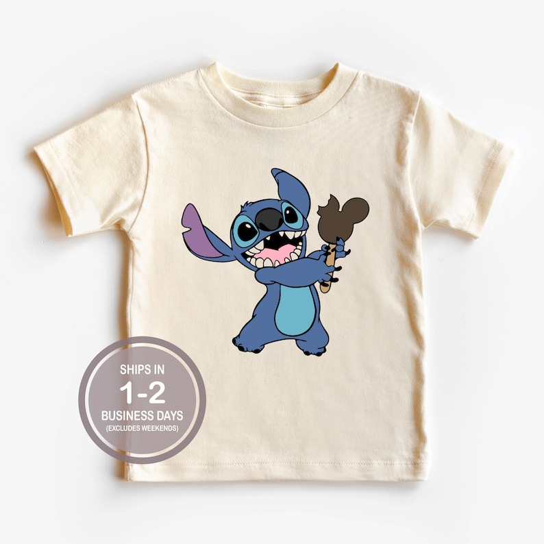 May include: A light beige t-shirt with a blue cartoon character holding a brown Mickey Mouse shaped ice cream. The character has large blue eyes and a wide smile. The text "SHIPS IN 1-2 BUSINESS DAYS (EXCLUDES WEEKENDS)" is printed on the bottom of the shirt.