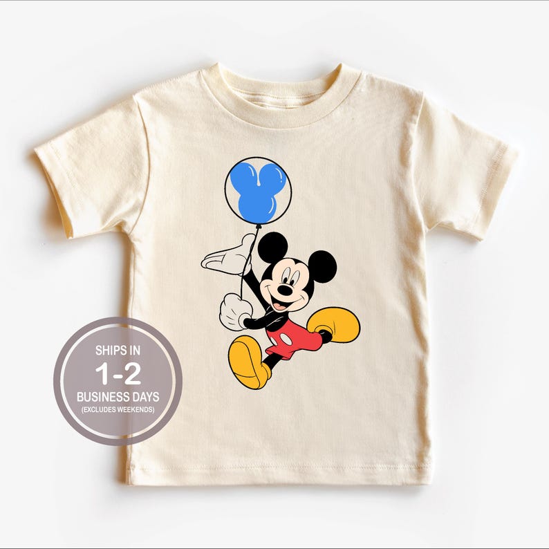 Puede incluir: Una camiseta beige de manga corta con una imagen de dibujos animados de Mickey Mouse sosteniendo un globo azul con un dise&ntilde;o de cabeza de Mickey Mouse. El texto "SHIPS IN 1-2 BUSINESS DAYS (EXCLUDES WEEKENDS)" est&aacute; impreso en la camiseta.