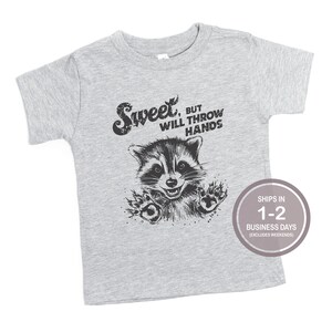 以下が含まれることがあります： グレーの半袖Tシャツに、黒と白のラクーンドローイングと「Sweet, but will throw hands」のテキストがプリントされています。